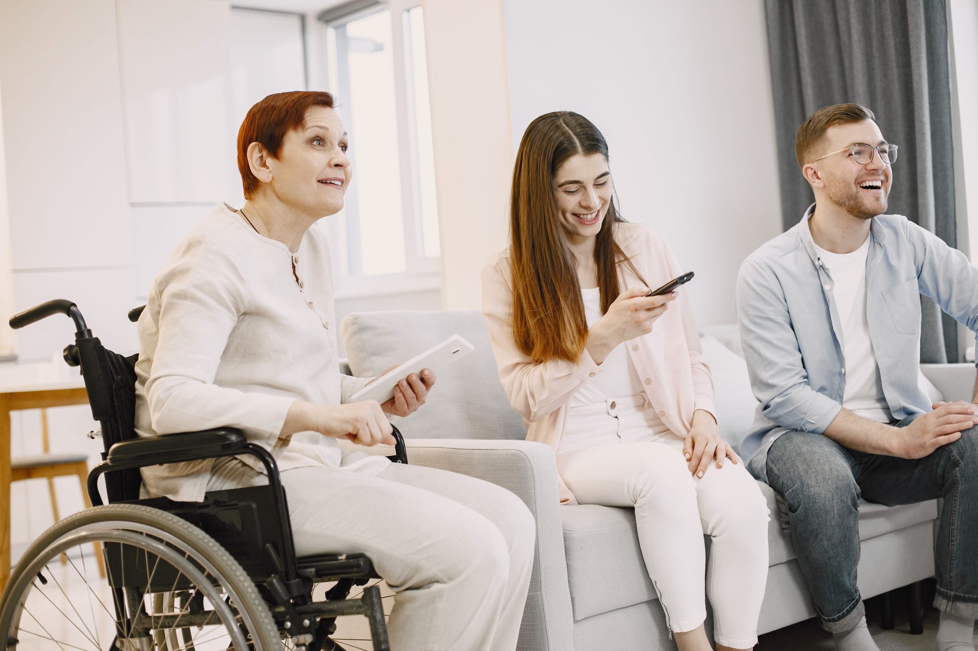 Friends laughing together on a couch at home, one using a wheelchair